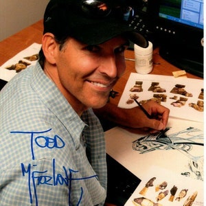 以下が含まれることがあります： 黒い野球帽とサングラスをかけた男性が白い紙に絵を描いています。彼は翼のある生き物を含むデザインに取り組んでいます。紙は他の紙や道具のあるテーブルの上に置かれています。男性の署名「Todd McFarlane」は、紙に青いインクで書かれています。