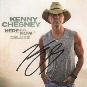 May include: Album cover for Kenny Chesney's "Here and Now Deluxe" featuring a man wearing a straw cowboy hat and a gray sleeveless shirt. The album title and artist name are printed in green and white. A signature is visible.
