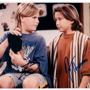 May include: A photograph featuring two young people, each with light brown hair. One is holding a black object, wearing a blue t-shirt with a basketball graphic. The other wears a brown shirt with a striped vest. Both have signatures.