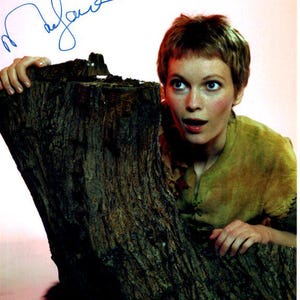 May include: A woman with short brown hair is looking at the camera with a surprised expression. She is wearing a brown shirt and is leaning against a tree trunk. The signature "Melanie" is written in blue ink at the top of the image.