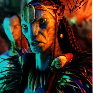 May include: A blue-skinned alien with long black braids and a feathered headdress. The alien has a large, round, white ornament on its forehead. The alien is looking directly at the camera with a serious expression. The image is signed "Wes Studi".