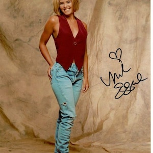 May include: A woman with short blonde hair is wearing a red sleeveless vest and blue jeans with a distressed look. She is standing in front of a brown backdrop and has signed her name "Jill Zarin" with a heart symbol above it.