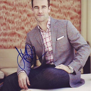 May include: A man wearing a grey plaid blazer over a pink and white checked shirt, blue jeans, and a watch. He is sitting on a white surface and has signed his name in blue ink.