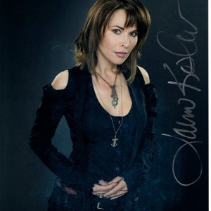 Könnte beinhalten: Eine Frau mit braunen Haaren trägt ein schwarzes Oberteil mit Ausschnitten an den Schultern und eine silberne Halskette mit einem Anhänger. Sie steht vor einem dunklen Hintergrund und hat ihre Hände vor sich gefaltet. Das Bild ist signiert mit "Alana Hael"