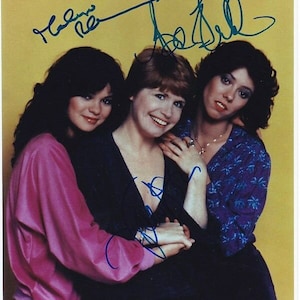 May include: A signed photograph of three women, two with dark hair and one with red hair, smiling and posing together. The women are wearing casual clothing. The photograph is signed by all three women.