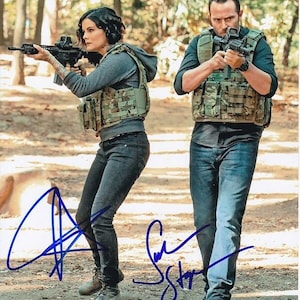 Puede incluir: Dos personas con equipo táctico sosteniendo rifles, posando al aire libre. Llevan chalecos de camuflaje, pantalones oscuros y botas. La imagen está firmada con tinta azul.