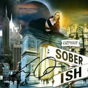 Puede incluir: Una mujer con un top rojo y pantalones negros posa frente a un telón de fondo de la ciudad de Nueva York. La imagen presenta el icónico Arco de Triunfo y señales de tráfico que dicen "Lizphair" y "Sober Ish".