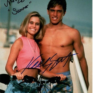 May include: A couple poses on a beach with a surfboard and a bicycle. The woman is wearing a pink tank top and denim shorts. The man is shirtless and wearing blue and white patterned swim trunks. The image is signed by both individuals with the text "Melissa" and "Summer" and "Kelly Slater".