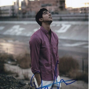 Op de afbeelding: Een man in een roze overhemd met knopen en een witte broek staat buiten. Hij kijkt naar de lucht. De achtergrond is wazig en toont een skyline.