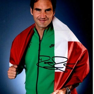 May include: A man wearing a green jacket with a Nike logo and a white and red flag draped over his shoulders. He is smiling and holding the flag with his right hand. The flag has a white stripe in the middle and red stripes on either side. The man is holding the flag in front of a blue background. The man's signature is visible on the flag.