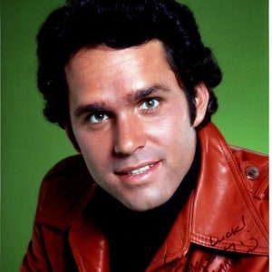 May include: A studio portrait of a man with dark hair, wearing a red leather jacket over a black turtleneck. The jacket is signed with a message. The background is a solid green.