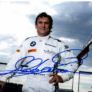 May include: A man wearing a white racing suit with blue and red accents. The suit has the text "Real Motorsport" and "Blancpain GT Series" on the chest. The man is signing his name in blue ink.