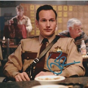 May include: A man wearing a brown uniform shirt with a badge and a brown tie sits at a table with a plate of food in front of him. He has a serious expression on his face.