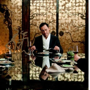 May include: A man in a black suit and white shirt sits at a table in a dimly lit room. He is eating from a plate with chopsticks. The table is set with glasses of wine, plates, and a teapot. The room has a dark wood floor and walls with a gold and brown patterned wallpaper.