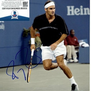 May include: A black and white photo of a tennis player in mid-swing. The player is wearing a black shirt, white shorts, and a white headband. The player is holding a tennis racket in their right hand. The player is on a tennis court with a blue surface. The player is signed by Roger Federer. The image includes the text "Hein" and "CERTIFICATION NUMBER: BB13033".