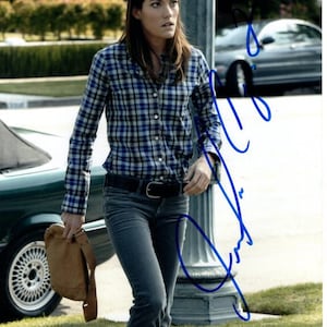 May include: A woman wearing a blue and white plaid shirt, dark blue jeans, and a brown leather bag. She is standing on a sidewalk in front of a green car and a gray car.