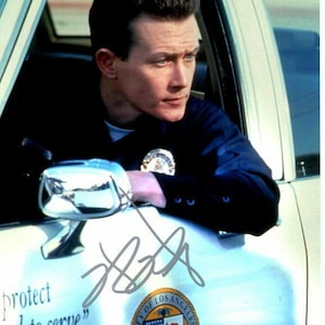 May include: A black and white photo of a man in a police uniform, sitting in a white car. He is looking to the right, and his hand is resting on the car door. The car has the words "protect and to serve" written on the side, along with the City of Los Angeles seal. The man's signature is visible on the car door.