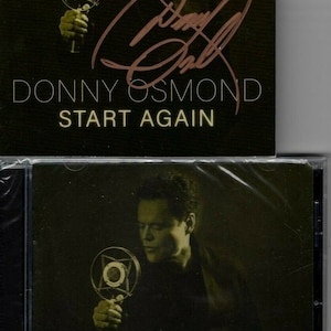 Puede incluir: Una caja de CD negra con fondo dorado y una imagen de Donny Osmond sosteniendo un micrófono. El texto "Donny Osmond Start Again" está impreso en la parte delantera de la caja.