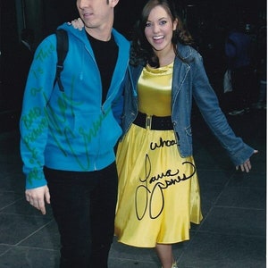 Peut inclure: Photographie de deux personnes, un homme et une femme, debout côte à côte. L'homme porte un sweat à capuche bleu et un pantalon noir. La femme porte une robe jaune, une veste en jean et des chaussures blanches. Les deux ont des autographes sur leurs vêtements.