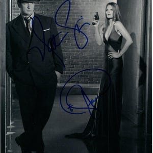 May include: Monochrome photograph of a man and a woman in a room. The man is in a dark suit and fedora. The woman holds a gun and wears a long, dark dress. Both figures have blue ink signatures on the image.
