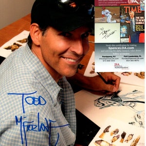 以下が含まれることがあります： 男性が手のイラストにサインをしているクローズアップ。男性は、格子縞のシャツと黒い野球帽をかぶっています。サインは「Todd McFarlane」とあります。画像には、TT48852という番号のJSA認証ステッカーも含まれています。