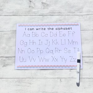 May include: A white dry-erase board with a pink and blue wavy border. The board has dotted lines for tracing the alphabet, with the letters "Aa Bb Cc Dd Ee Ff Gg Hh Ii Jj Kk Ll Mm Nn Oo Pp Qq Rr Ss Tt Uu Vv Ww Xx Yy Zz" printed in black. A white dry-erase marker is next to the board.