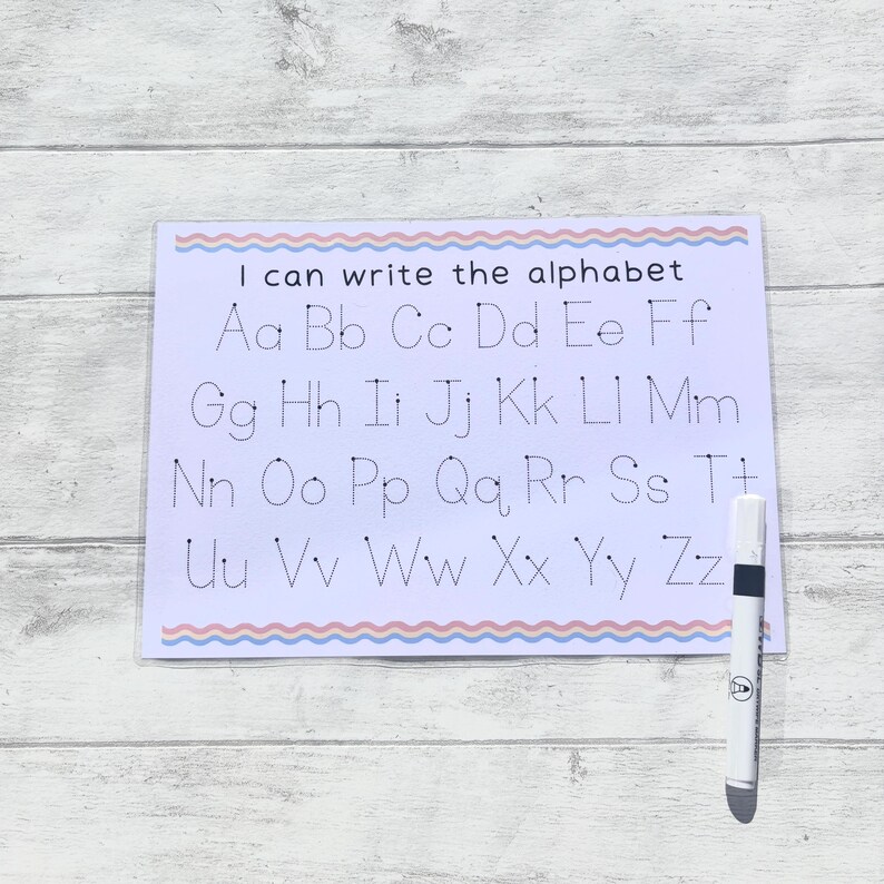 May include: A white dry-erase board with a pink and blue wavy border. The board has dotted lines for tracing the alphabet, with the letters "Aa Bb Cc Dd Ee Ff Gg Hh Ii Jj Kk Ll Mm Nn Oo Pp Qq Rr Ss Tt Uu Vv Ww Xx Yy Zz" printed in black. A white dry-erase marker is next to the board.