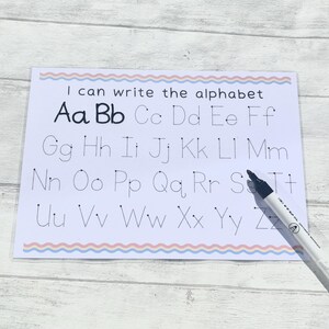 May include: A white sheet of paper with a blue and pink border. The paper has dotted lines for tracing the alphabet, with the letters "Aa Bb Cc Dd Ee Ff Gg Hh Ii Jj Kk Ll Mm Nn Oo Pp Qq Rr Ss Tt Uu Vv Ww Xx Yy Zz" printed in black. The text "I can write the alphabet" is printed at the top of the sheet.