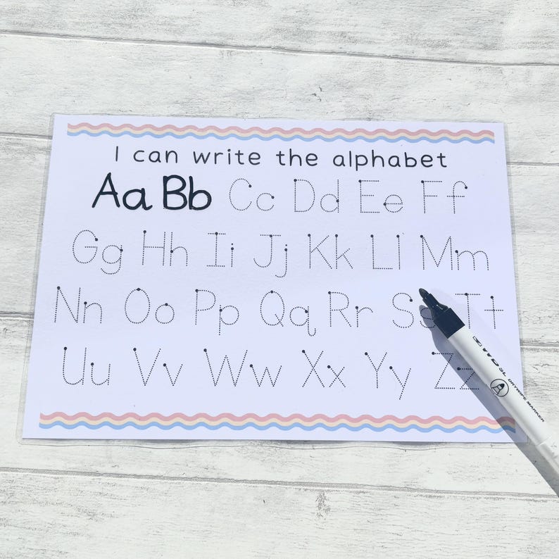 May include: A white sheet of paper with a blue and pink border. The paper has dotted lines for tracing the alphabet, with the letters "Aa Bb Cc Dd Ee Ff Gg Hh Ii Jj Kk Ll Mm Nn Oo Pp Qq Rr Ss Tt Uu Vv Ww Xx Yy Zz" printed in black. The text "I can write the alphabet" is printed at the top of the sheet.
