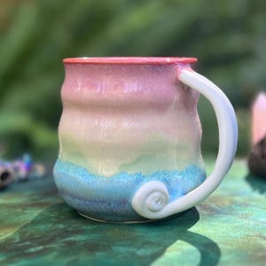 May include: A handmade ceramic mug with a pink, yellow and blue glaze. The mug has a unique, wavy design and a white handle with a swirl detail.