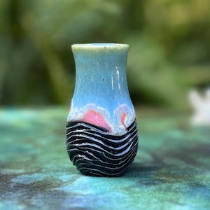 May include: A small, blue and black ceramic vase with a pink and white design. The vase is sitting on a green and blue surface.