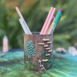 May include: A brown pen holder with a wood-grain texture and green and yellow mushroom and plant details. The holder contains several pencils and pens. The holder is cylindrical and sits on a green surface.