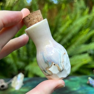 May include: A small, pearlescent white ceramic bottle with a cork stopper. The bottle has a textured, faceted design on the lower half and iridescent blue and gold accents. The bottle is held against a backdrop of green foliage.