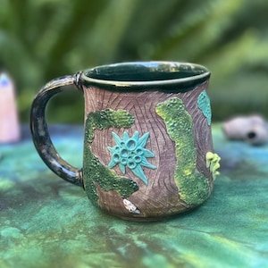 May include: A handmade ceramic mug with a brown, textured exterior resembling tree bark. The mug features green and turquoise accents, including a floral design and moss-like embellishments. The handle and rim are dark green.