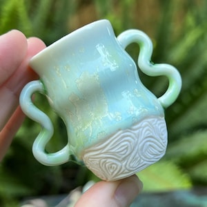 May include: A small, white ceramic cup with two handles. The cup has a light blue glaze with a white design on the bottom. The handles are green.