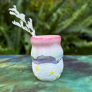 May include: A small, handmade ceramic vase with a colourful design. The top is pink and fades into blue and green. The bottom has a white, textured pattern with yellow star accents. A sprig of white dried flowers is inside.