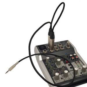 May include: A black audio cable with two 1/4 inch male connectors plugged into a Behringer Xenyx 502 USB audio mixer. The mixer has a variety of knobs and buttons for controlling audio levels and effects.