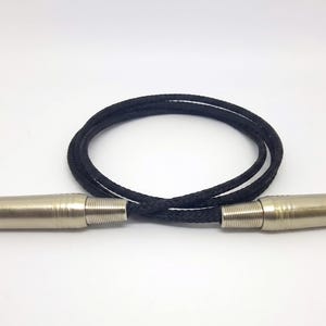 May include: A black braided audio cable featuring two silver-toned metal 1/4-inch TRS jack connectors at each end. The cable is coiled into a circular shape, showcasing the textured fabric exterior of the cord and the metallic finish of the plugs.