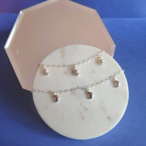 May include: A silver chain anklet with multiple crescent moon charms hanging from it. The anklet is displayed on a white marble surface.