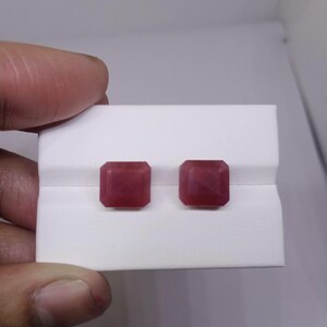Puede incluir: Dos gemas cuadradas de color rojo intenso se exhiben sobre una superficie blanca. Las gemas son sostenidas por una mano, mostrando sus facetas pulidas y su rico color. El entorno es sencillo, enfatizando la belleza de las piedras.