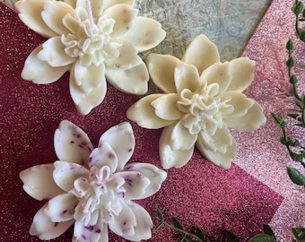 Puede incluir: Tres velas de cera blanca en forma de flor con un diseño detallado. Las velas están sobre una superficie rosa brillante.