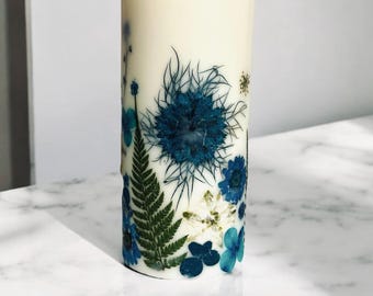 Peut inclure: Une bougie cylindrique avec un extérieur blanc crème, ornée de fleurs bleues pressées et de feuilles de fougère vertes. L'arrangement floral est disposé autour de la base de la bougie, créant une esthétique naturelle et décorative.