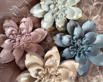 May include: Four decorative flower-shaped objects in shades of pale blue, pink, and cream. The flowers have detailed petal designs and are arranged on a lace surface. The objects appear to be made of a solid material.