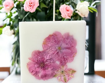 Peut inclure: Une bougie carrée blanche avec trois fleurs roses pressées. La bougie a une mèche blanche.