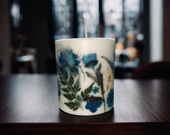 Può includere: Una candela bianca con un design floreale. La candela ha fiori e foglie blu e verdi pressati incorporati nella cera.