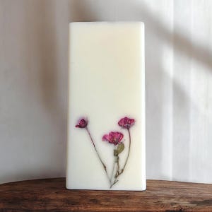 May include: A rectangular, off-white candle with three pink flowers embedded in the wax. The flowers have delicate petals and green stems. The candle is displayed on a wooden surface.