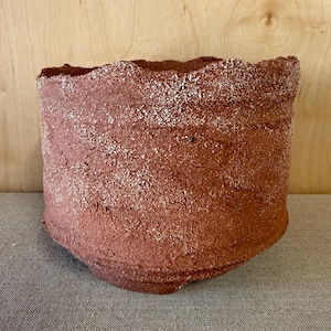 May include: A cylindrical ceramic vessel with a deep terracotta red hue and a heavily textured, granular surface. The rim is uneven and rugged, featuring a distinct contrast where white, crystalline particles are embedded into the clay body.