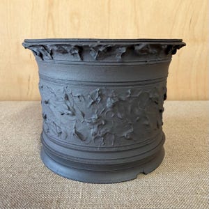 May include: A matte black ceramic planter with a cylindrical shape, featuring a textured, rugged surface pattern around the center and a smooth, flared rim at the top. The base is flat and circular, providing a stable foundation for the vessel.