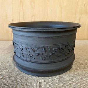 May include: A matte black ceramic bowl with a wide, circular opening and a thick, flared rim. The exterior features a prominent, textured band around the center with irregular, crinkled, and raised clay details, contrasting with the smooth base.