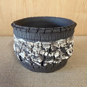May include: A round ceramic bowl with a dark charcoal gray base and a wide, textured band around the center featuring white, cracked, bark-like detailing. The rim and lower section exhibit a deep, matte black finish with vertical fissures.